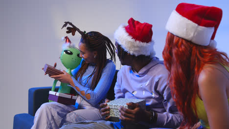 Studio-Shot-Of-Gen-Z-Friends-Giving-Presents-For-Christmas-Sitting-On-Sofa-Wearing-Santa-Hat-And-Reindeer-Antlers-1